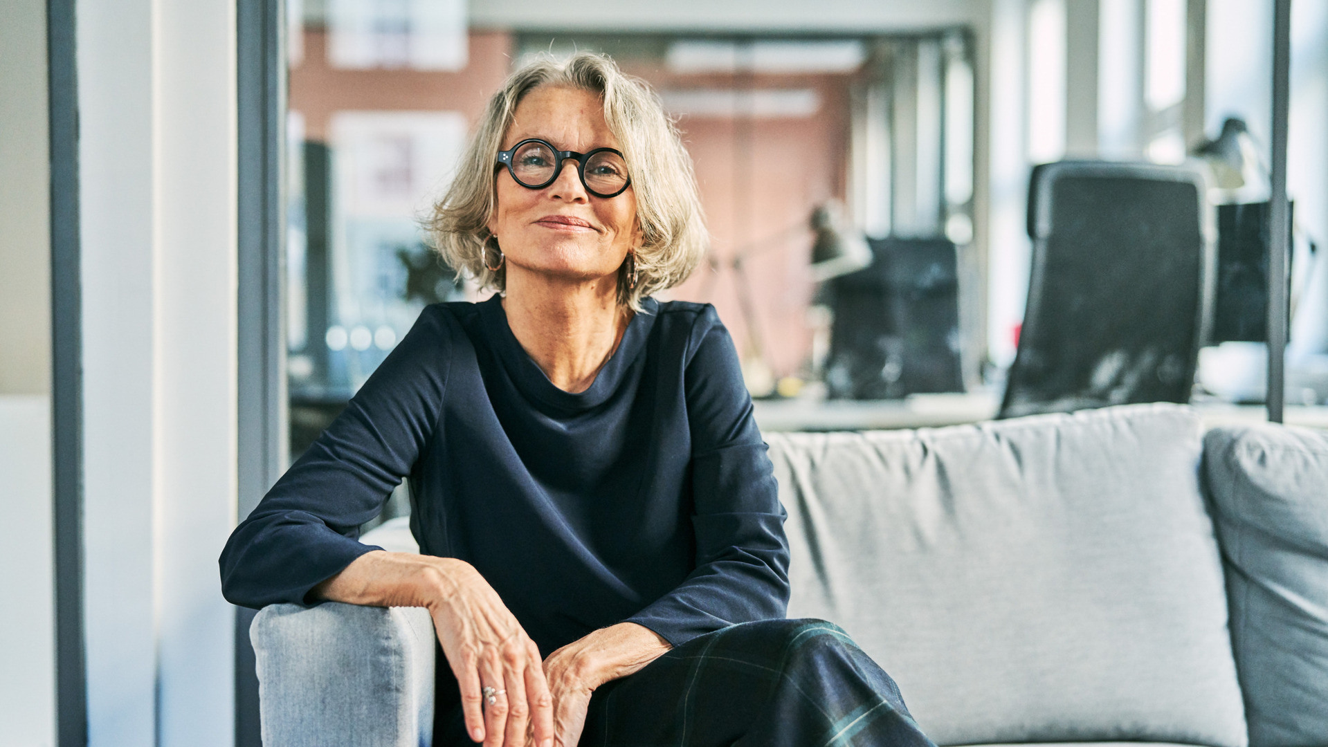 Kvinne med kort krøllete hår og briller sitter i en grå sofa og smiler mot kamera