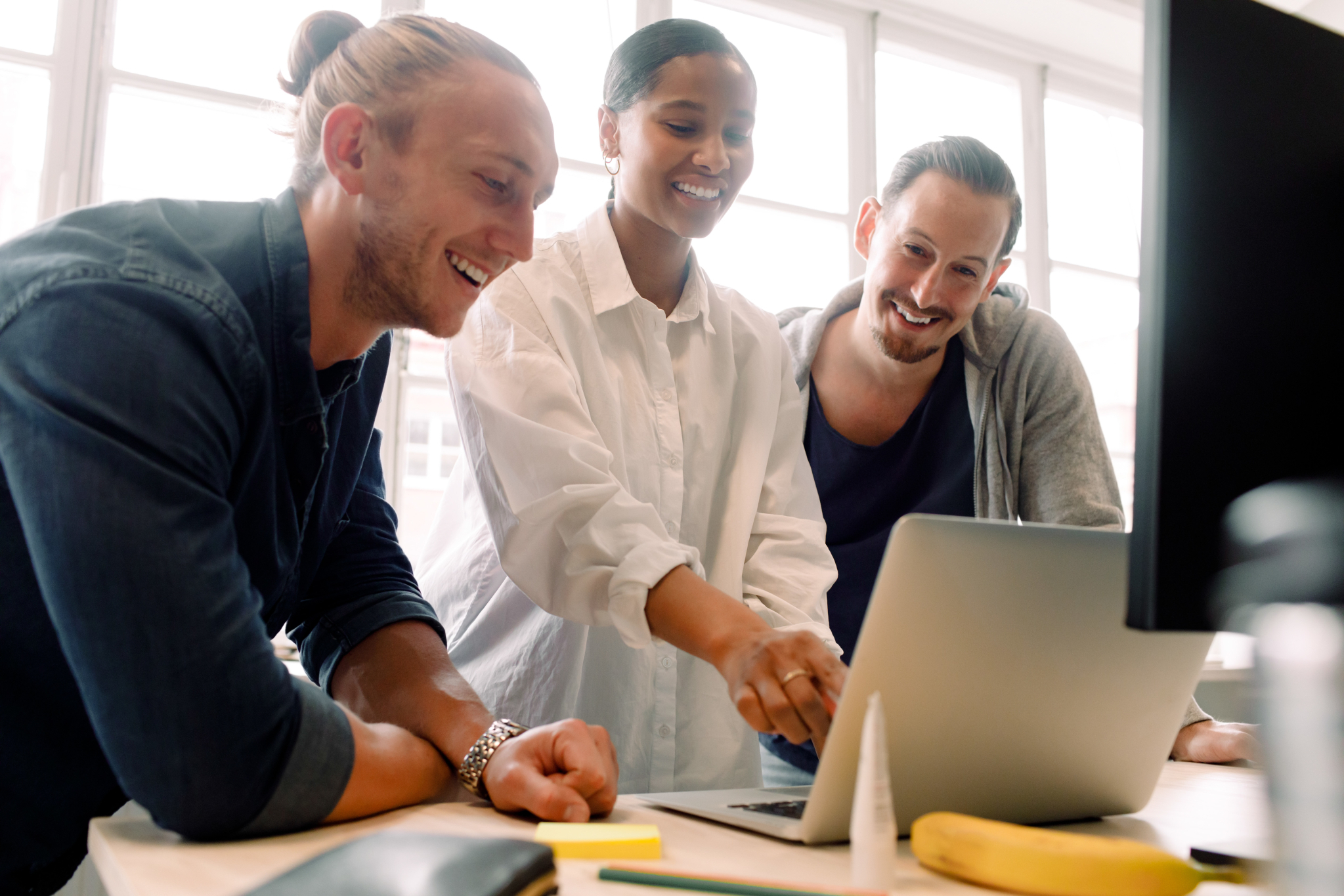 Tre unge mennesker står rundt en stor skjerm og samarbeider om noe på skjermen, de smiler og ler.