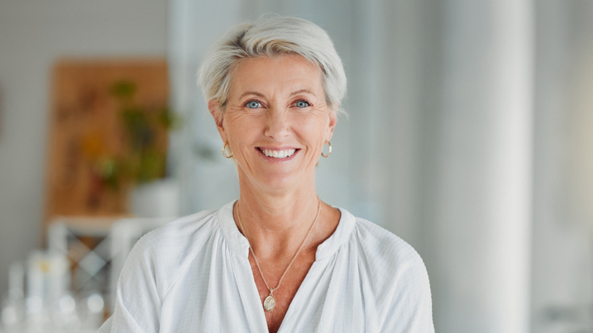 Kvinne med kort grått hår og hvit skjorte står med hendene i kors og ser mot kamera