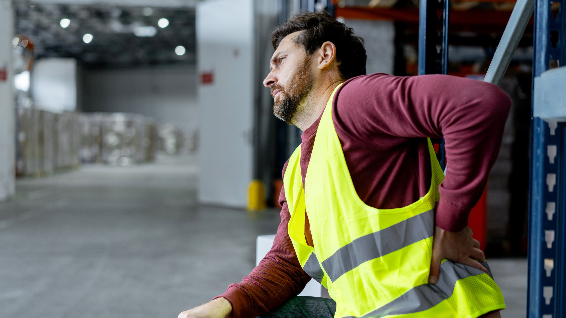 Mann med gul vest sitter på et lager og holder seg til ryggen i smerte