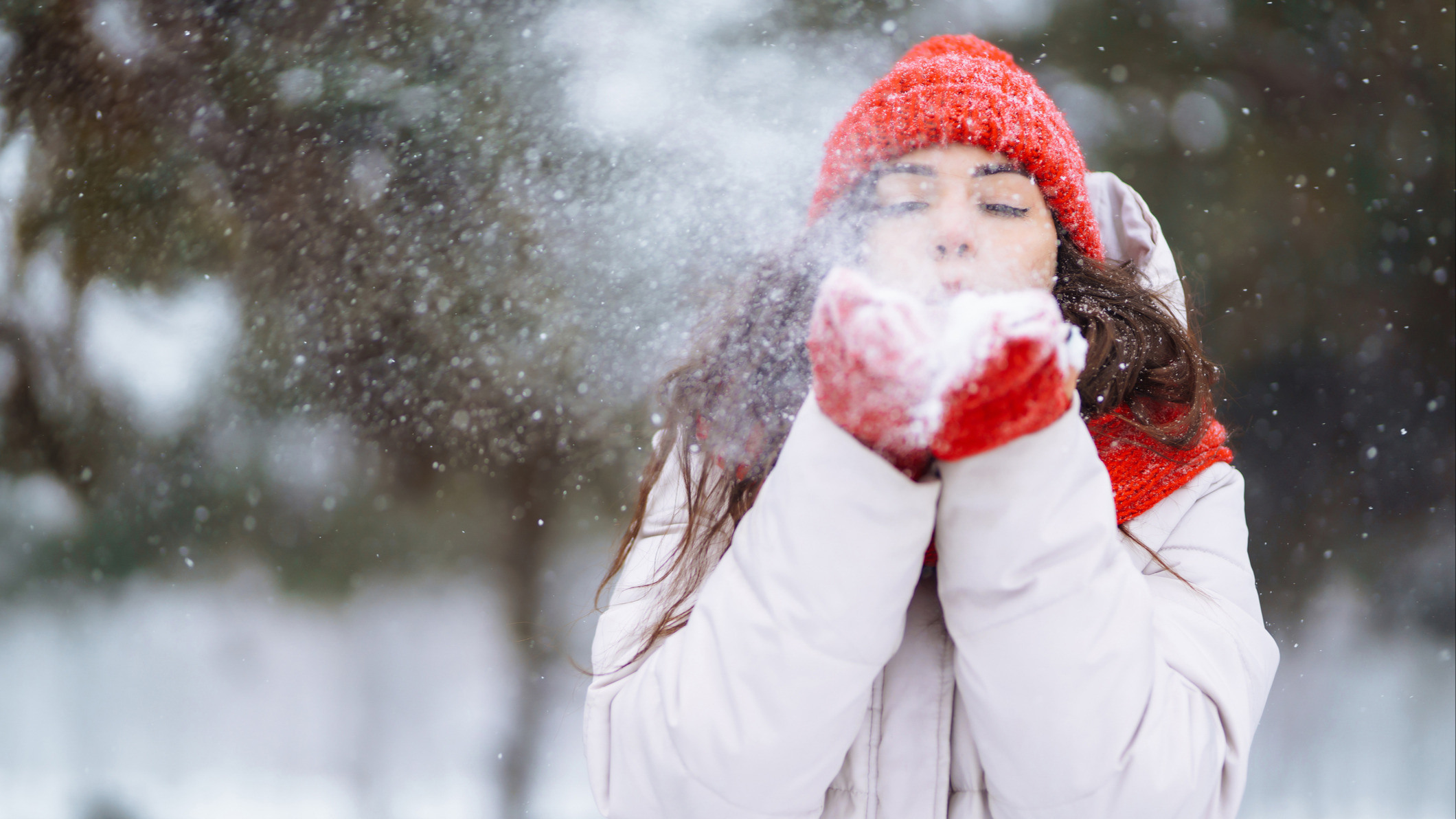Kvinne i rød lue og røde votter som blåser snø mot kamera.